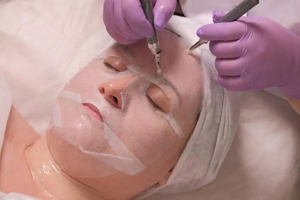 Microcorriente alisado de arrugas. Un cosmetólogo profesional en guantes lila trabaja con dos electrodos a través de una máscara en la frente de una mujer. Procedimiento cosmético rejuvenecedor. Levantamiento de piel — Foto de Stock