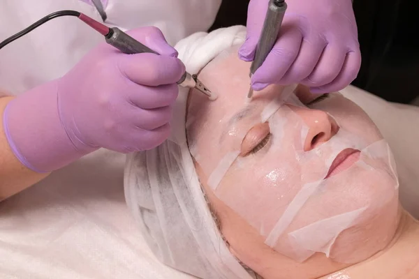 Levantamiento. Microcorriente alisado de arrugas. Un cosmetólogo profesional en guantes lila trabaja con dos electrodos a través de una máscara en la frente de una mujer. Procedimiento de rejuvenecimiento cosmético . — Foto de Stock