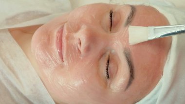 Spa'daki kanepede yatan beyaz havlulu güzel bir kızın yakın çekim. Güzellik uzmanı bir fırça ile yüzünde bir kadın krem uygulanır. Yüz güzellik tedavileri