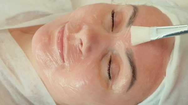 Spa'daki kanepede yatan beyaz havlulu güzel bir kızın yakın çekim. Güzellik uzmanı bir fırça ile yüzünde bir kadın krem uygulanır. Yüz güzellik tedavileri