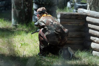 Ormanda Paintball oyunu. Yaz. Elinde silah olan kamuflaj kıyafetleri giyen bir kız ahşap bir barınağın arkasından bakıyor..