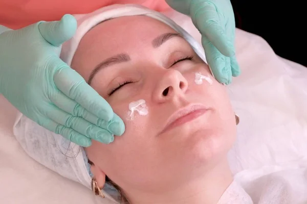 Esteticista en guantes aplicar crema hidratante en las mejillas de la chica. Una buena mujer toma un masaje facial. Foco suave. Sala de tratamiento de belleza para el cuidado facial y corporal . — Foto de Stock