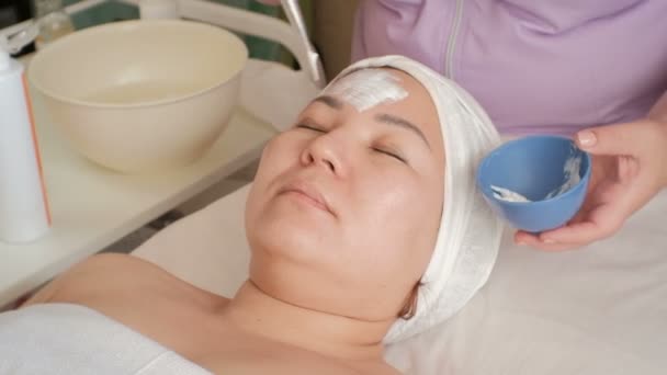 Asian Girl w salonie piękności. Kosmetyczka stosuje biały krem do masażu na twarz muzułmańskiej kobiety z pędzlem. Przyspieszone wideo. Upływ czasu. Zabieg kosmetyczny do oczyszczania skóry. — Wideo stockowe