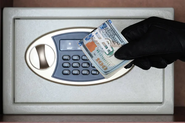 A black glove holds a wad of American dollars in front of a safe with a combination lock. The concept of protecting money and jewelry from theft. Keeping money in a safe place