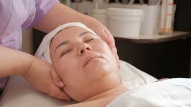 Asyalı bir kız için yüz masajı. Bir güzellik uzmanının ellerinin yakın çekim, bir kadın müşteri başının yan dönüyor. Güzellik salonunda terapötik prosedür.