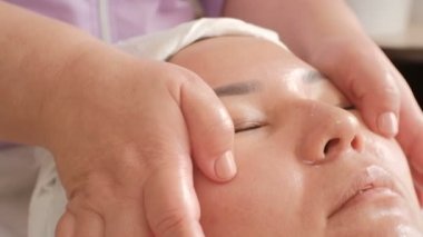 Anti-aging yüz masajı. Close. Profesyonel bir kozmetolojistin elleri Asyalı bir kızın yüzündeki deriyi pürüzsüzleştirin. Spa güzellik salonu.