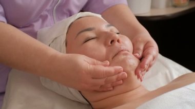 Anti-aging yüz masajı. Profesyonel bir kozmetolojistin elleri Asyalı bir kızın yüzündeki deriyi pürüzsüzleştirin. Spa güzellik salonu.