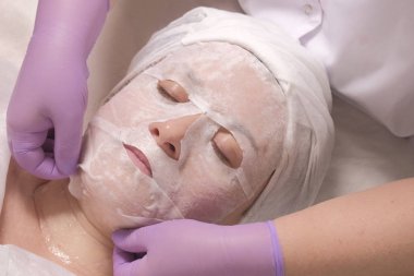 Gözleri kapalı bir kadın. Pembe eldivenli kozmetolojistin elleri, beyaz besleyici maskeyi kadın yüzünden çıkarır. Cilt tazeleme, sağlık ve güzellik kavramı. Profesyonel kozmetoloji.