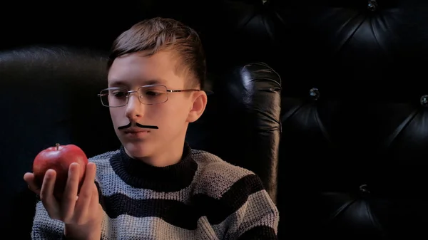 A teenager in glasses with a glued-on mustache, wearing a sweater, sits in a black leather chair and looks thoughtfully at the red apple in his hand. Dark background. — Stock Photo, Image