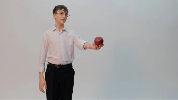 A teenager in glasses, wearing a white shirt and black pants, holds an apple in his hands and stretches it forward. Light background. Copy space. Back to school. — Stock Photo, Image