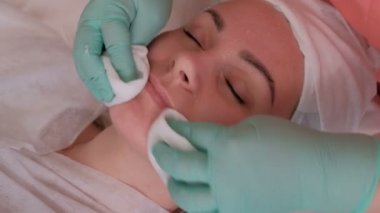 Bir güzellik salonunda gençleştirici bir prosedür kapalı gözleri ile kız. Eldivenli bir güzellik uzmanının elleri. Profesyonel bir kozmetolojist bir kadın yüzünden kozmetik jel kalıntılarını siler..