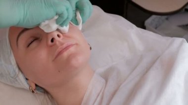 Profesyonel bir kozmetolojist-terapist bir kadın yüzünde şeffaf bir kozmetik jel kalıntıları siler. Bir güzellik salonunda bir prosedür kapalı gözleri ile kız. Eldivenli bir güzellik uzmanının elleri.