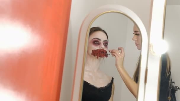 Die Spiegelung im Spiegel. Zwei Mädchen bereiten sich auf Halloween vor. Make-up-Bild einer furchterregenden Dame mit einem zerlumpten blutigen Mund. Erstellen eines Kostüms für eine Horrorparty oder Allerheiligen. — Stockvideo