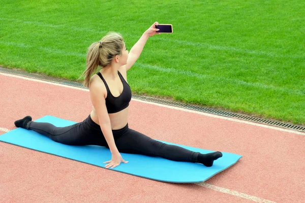 Una ragazza si fa un selfie mentre è seduta su uno spago su un tappeto blu. Ginnastica, yoga, stretching le gambe all'aria aperta accanto all'erba verde. Stile di vita sano . — Foto Stock