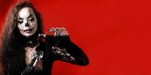 A Halloween-style brunette wearing black clothes pours a red liquid that looks like blood into a skull-shaped glass dish. A blank for invitation card, banner or poster on the theme of a terrible party — Stock Photo, Image