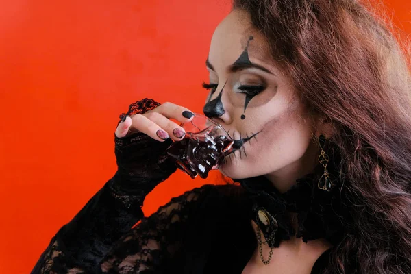 Halloween makeup. A brunette girl with long dark hair in a black dress drinks a bloody liquid from a glass in the form of a skull. Copy space, red background. Concept. The feast of a costume carnival. — Stock Photo, Image
