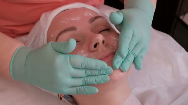 戴着绿色手套的美容师的手 在女人的脸上滋润滋润滋润的奶油 在美容院的手术过程中 眼睛闭着眼睛的女孩闭着笑容 — 图库视频影像