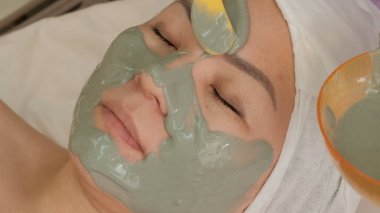 Gençleştirici bir prosedür bir güzellik salonunda Asyalı orta yaşlı kadın. Bir kadın yüzünün yakın çekim. Güzellik kız istemcinin cilt üzerinde gri bir aljinat maskesi uygular. Yaşam tarzı, sağlık, güzellik.
