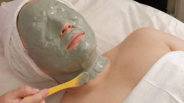 Kadın yüzü tamamen bir kozmetik maske ile kaplıdır. Güzellik kızın cildine gri bir kitle uygular. Gençleştirici bir prosedür bir güzellik salonunda orta yaşlı kadın.