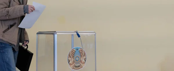 Qazaqstan, 6-9-2019, Elections. A girl in a jacket with a briefcase in the voting room wants to put the ballot in a transparent box with the coat of arms of Kazakhstan. Copy space. Blank for banner. — Stock Photo, Image