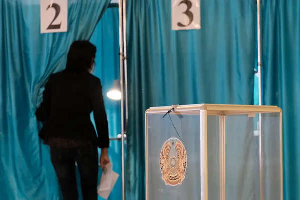 Kazakhstan, Qazaqstan, June 9, 2019, Elections, Voting room. A woman walks into an individual booth with numbers two. Transparent box with the emblem of Kazakhstan for counting votes. Blue background. — Stock Photo, Image