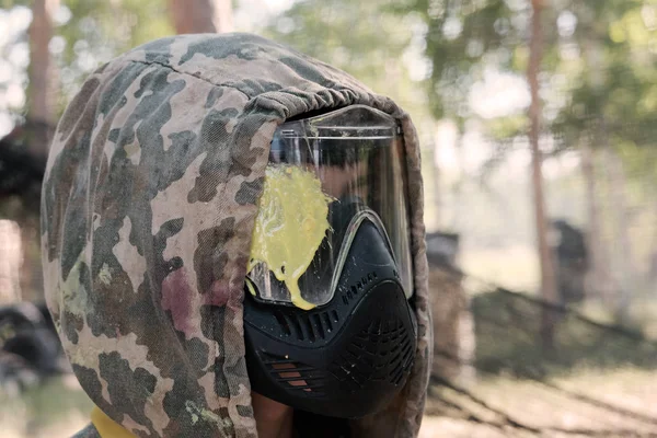 Headshot of a paintball player. The guy in the profile in a camouflage hood in a protective mask with a yellow blot on the glass in the forest. Sports lifestyle or entertainment and team war games. — Stock Photo, Image