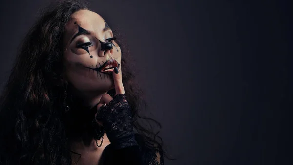 Makeup for Halloween. Sexy brunette girl in black clothes with a scary art on her face keeps her index finger on her lips. Copy space. Advertising concept for All Saints' Day or for the devil's party. — Stock Photo, Image