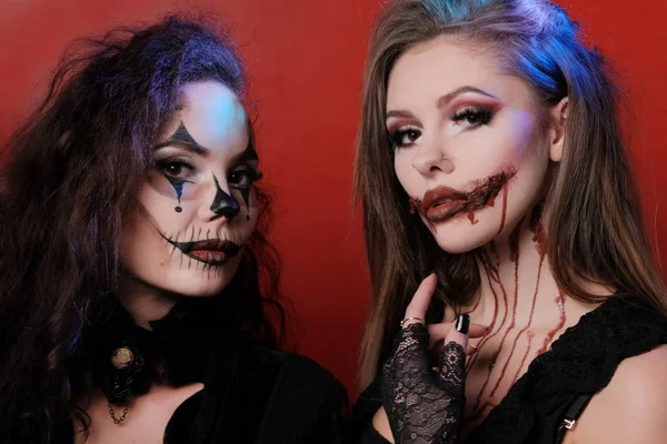 Portrait of two beautiful sexy girls in black clothes with scary bodypainting on their faces. Makeup for Halloween. Red background. All Saints' Day. Festive party in the devilish style. — Stock Photo, Image