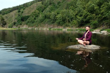 Uzun kollu bir gömlek bir kakvkazskoy görünümü ile bir adam bir nehir veya gölde bir taş üzerinde oturur. Ormanda yoga kavramı, dağlarda gevşeme, seyahat ve yaşam tarzı. Kopyalama alanı..