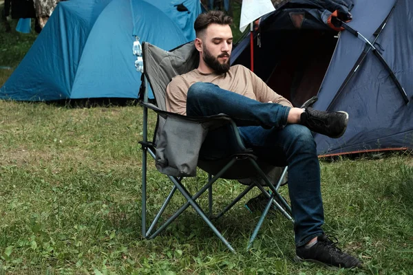 A srác egy szakállas farmer és egy póló ül egy összecsukható szék közelében a sátrak. A koncepció az Mans szabadidő a természetben, az utazás és életmód, hegyi turizmus. — Stock Fotó