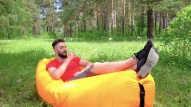 Kırmızı tişörtlü adam ormandaki yeşil çimenlerin üzerinde şişme bir şilte üzerinde yatıyor. Bir adamın elinde bir kadeh şarap. Turist piknikte şarap içer. Konsept seyahat ve yaşam tarzı.