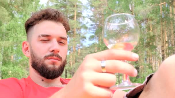 手里拿着一杯酒 留着胡须的布鲁内特在野餐时休息和喝酒 那个穿红色T恤的家伙躺在森林的空地上 快乐和生活方式的概念 — 图库视频影像