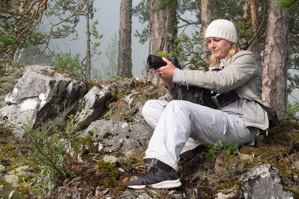 Hermosa chica en ropa blanca tomando fotos de la naturaleza. Un fotógrafo con sombrero y chaqueta sostiene una cámara profesional en sus manos y está sentado en una ladera de una montaña cerca de un árbol . — Foto de Stock