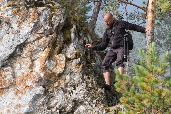 Fotógrafo viajante careca com uma barba desce da montanha segurando a árvore com a mão. Viagens e turismo. Nos ombros do saco caras com uma câmera e um tripé . — Fotografia de Stock