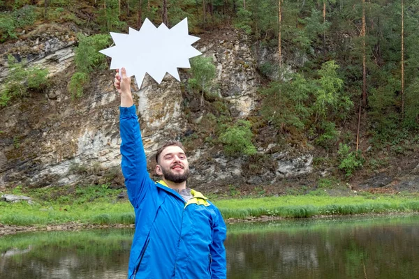 Descuentos de temporada para ropa de otoño y turismo. Un joven divertido con barba sonríe y sostiene un letrero blanco. El tipo de la chaqueta azul en la naturaleza en las montañas cerca del río . —  Fotos de Stock