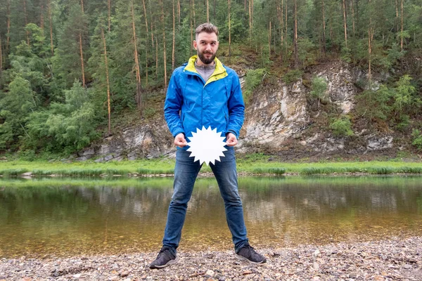 Temporada de descuentos para ropa de otoño para viajes. Un turista divertido con barba astuta mira a un lado y sostiene un plato blanco en forma de estrella en el nivel de la cintura. Un joven con una chaqueta azul. naturaleza — Foto de Stock