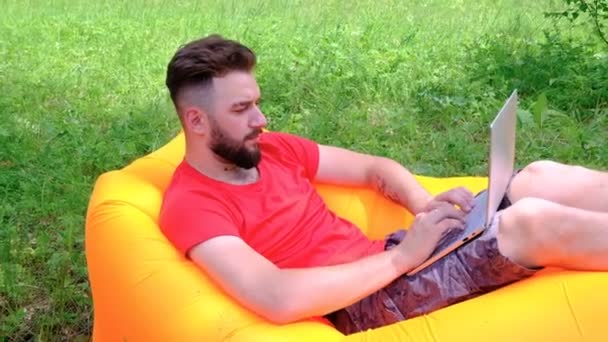 Ten chlápek v červeném tričku, ležící na Nafukovatelné matraci pracující na laptopu. Mladý muž s plnovousem je v přírodě nezávislý člověk. Koncepce volného času a podnikání, studium a životní styl. — Stock video