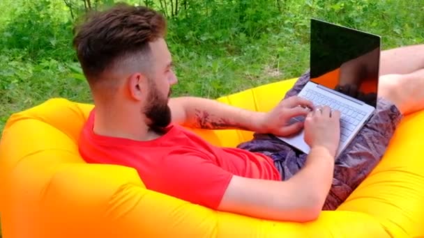 有胡子的商人 自由职业者或游戏者的性质 那个穿红色T恤的家伙躺在空中沙发上 在笔记本电脑上工作 休闲与商务 学习和生活方式的概念 — 图库视频影像