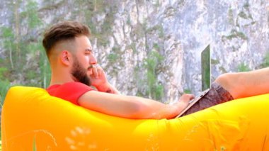 Şık adam işadamı serbest turuncu bir hava kanepe üzerinde bir dizüstü bilgisayar üzerinde çalışan ve bir cep telefonu konuşuyor yalan. Doğada dağların arka planında sakallı genç bir adam. Eğlence ve iş kavramı, çalışma ve yaşam tarzı.