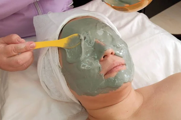 Grijze alginaat masker op het gezicht van een Aziatische vrouw. De handen van een schoonheidsspecialiste zetten veel op het gezicht van de cliënt. Tillen van de huid. Cosmetische verjongings procedure. Gesloten ogen. — Stockfoto