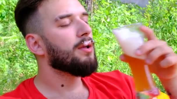 Le type au t-shirt rouge boit de la bière. Mousse sur la moustache. Une barbe de jeune homme se trouve sur un canapé d'air orange dans une clairière dans la forêt. Gros plan. Concept vacances, pique-nique, voyage et style de vie . — Video