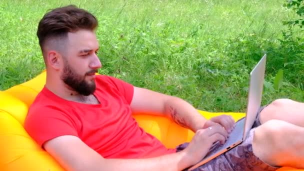 Un giovane uomo con una maglietta rossa è disteso su un divano ad aria e sta lavorando su un computer portatile. Il tizio con la barba fa affari online in un prato verde e soleggiato. Tempo libero, lavoro e viaggi. Concetto — Video Stock