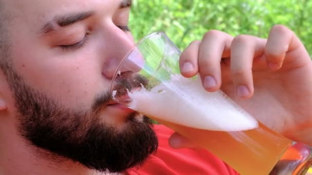 Egy fiatal férfi a piros póló italok sör örömmel. Közeli. A srác a szakálla ivott habos alkoholos ital ki egy pohár. Pihenés, piknik és utazás. Koncepció — Stock videók