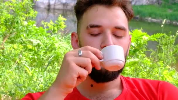 Un joven con barba disfruta bebiendo café en la naturaleza. Primer plano de un tipo brutal con una taza de té blanco en la mano contra el fondo de las montañas y un río . — Vídeos de Stock