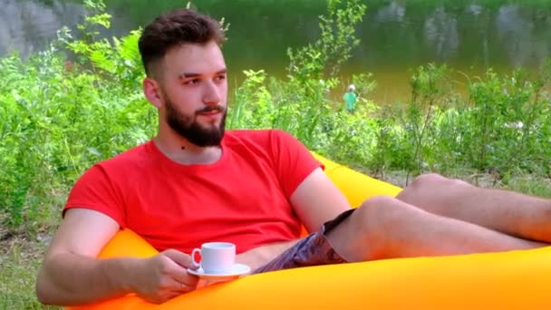 Młody mężczyzna w czerwonym T-shirt z filiżanką kawy leży na przewiewnej kanapie na tle rzeki. Facet z brodą trzyma białą filiżankę w ręku na górskiej, słonecznej łące. — Wideo stockowe