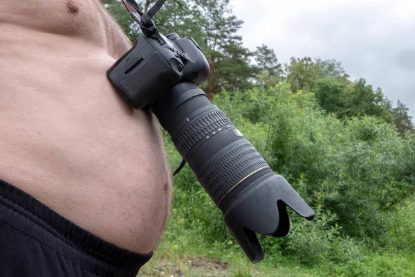 A digital camera with a large lens hangs around the neck of a big-bellied man. Curious frame landscape photographer. Close-up. Concept Forest background.
