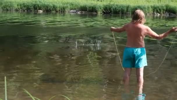 Pesca con mosca. El hombre adulto pesca el río de la montaña. Un pescador en pantalones cortos azules con la espalda desnuda arroja el cebo de pie hasta las rodillas en el agua. Deporte o hobby. Parece que Santas vacaciones de verano . — Vídeo de stock