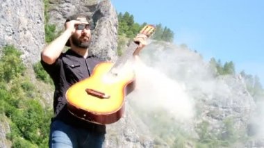 Siyah gömleğinde sakalı olan bir adam güneş gözlüğü koyar ve duman geldiği akustik bir gitar çalmak için davranır. Dağlarda doğanın bir arka planda bir müzisyen hakkında garip komik video.