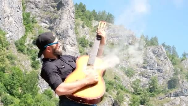 一个留着胡子 穿着黑色衬衫和太阳镜的家伙假装弹着一把烟熏的原声吉他 奇怪的有趣的视频 在山的自然背景的音乐视频 — 图库视频影像