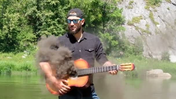 原声吉他发出黑烟 穿着衬衫 帽子和太阳镜的白种人男人假装在演奏音乐 一个奇怪的视频 关于嬉皮士的背景附近的河流在山中 — 图库视频影像